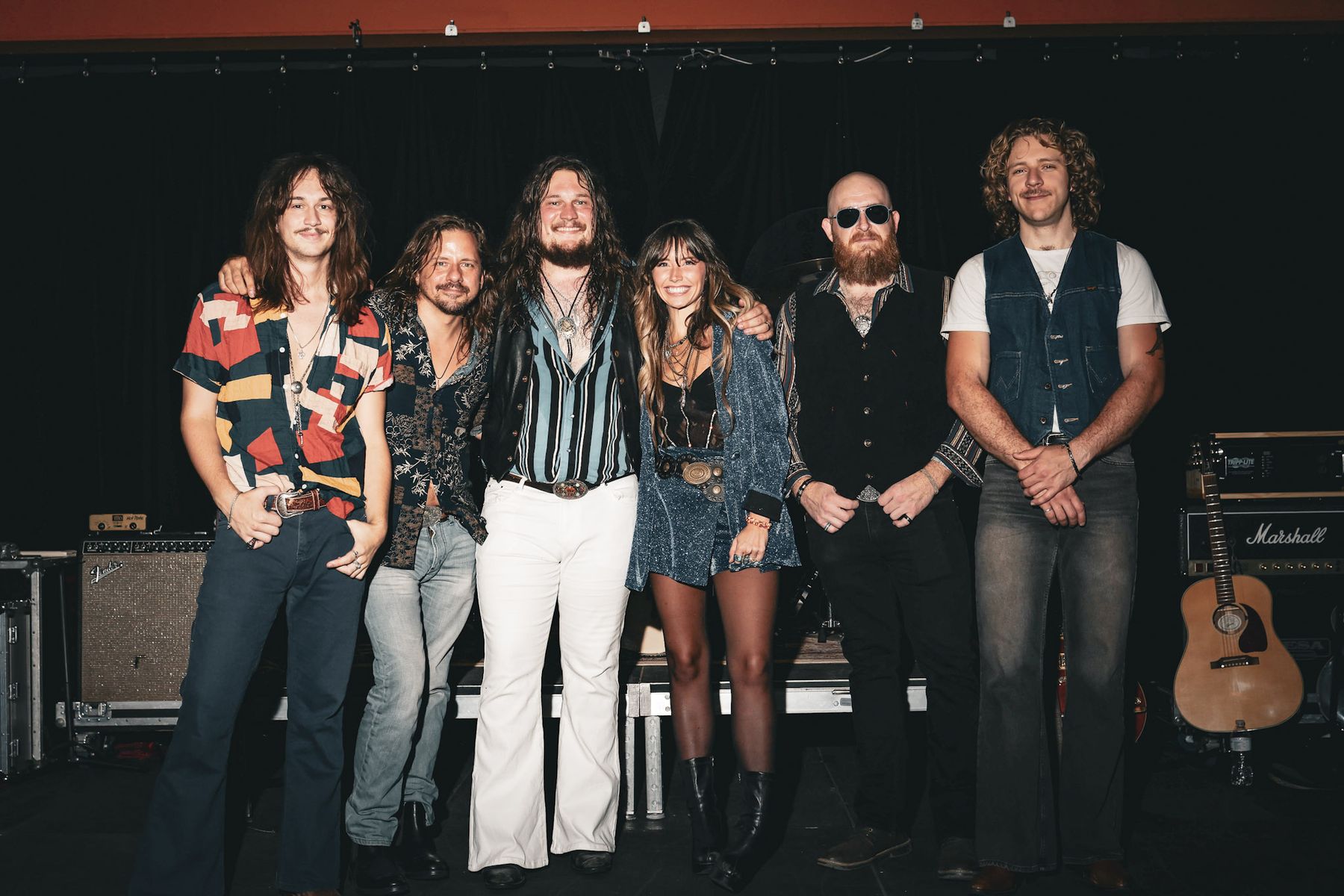 Band posed together on stage beside a guitar amp